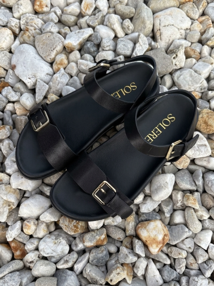 Black sandals with gold buckles on a pebble surface